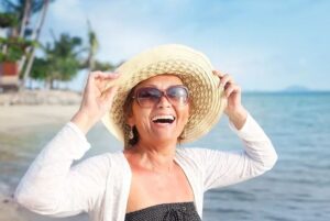 imagem de uma mulher com aproximadamente 55 anos com roupas de praia em um cenário paradisíaco. Ela usa óculos de sol e sorri levantando a aba do chapéu de sol.